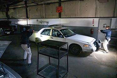 Two law enforcement academy students conduct a simulated vehicle stop during a tactical training exercise inside a training facility, using flashlights to assess a white patrol car in a low-light scenario.